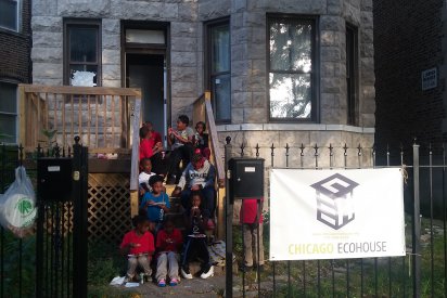 Participating youth sitting on the steps of Chicago Eco House. A sign on a wrought iron gage reads Chicago Eco House. Through the open gate entrance 10 children can be seen sitting on wooden steps up to a wooden deck off the the stone house, and beside its protruding bay window. The children are eating pizza.