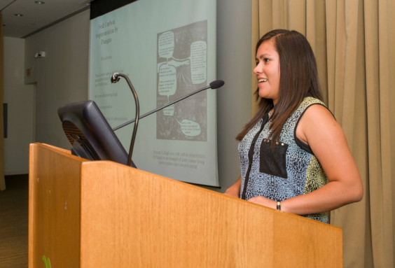 Erica Rocha standing at a podium. Podium and Erica face the left side of the photo, Erica standing on the right side. A powerpoint slide can be seen behind Erica, but the text cannot be made out.