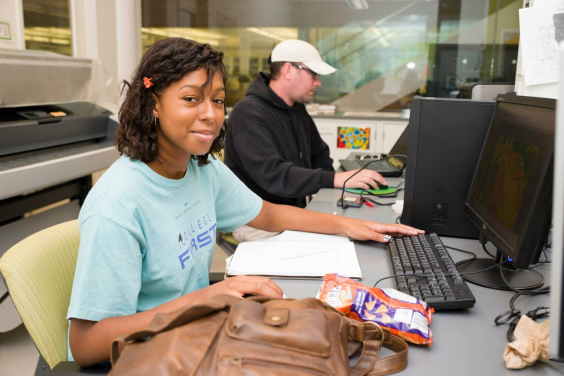Jannice Newson sitting at a computer. Jannice in on the left in the foreground, facing right and looking towards the photographer. A man in a baseball cap sits at another computer in the background.