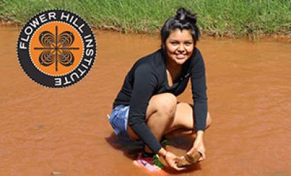 Teenage participant of E4 STEM kneeling in muddy water way with a grassy shore behind her. The Flower Hill Institute Logo brands the top left corner.