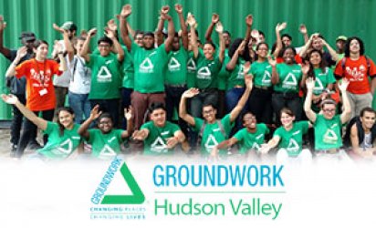 Green Team Greenway Corps members pose as a gropu in front of green cargo container and woods, many of the participants wearing green Groundwork Hudson Valley shirts. Many smiling faces and waving hands. A feathered white backdrop takes up the lower third with the Groundwork Hudson Valley logo over top in blue and green.