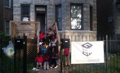 Participating youth sitting on the steps of Chicago Eco House. A sign on a wrought iron gage reads Chicago Eco House. Through the open gate entrance 10 children can be seen sitting on wooden steps up to a wooden deck off the the stone house, and beside its protruding bay window. The children are eating pizza.