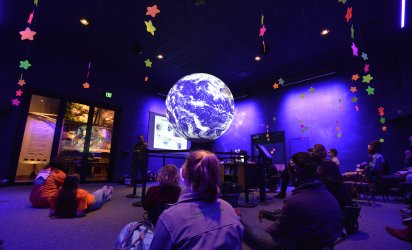 Purples and blues dominate the image. Kids sit in a dim light, lit in purple and blues, as a large globe takes center frame, in front of an adult speaker mid-presentation. Neon stars hang from the cieling.