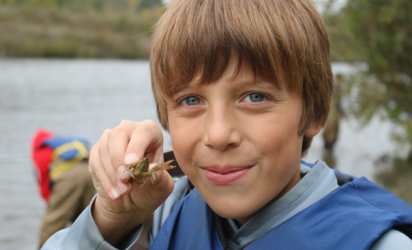 A NEMIGLSI student holds a crayfish up to the camera.  Behind the student a lake and trees can be seen out of focus, along with one person looking into the water.