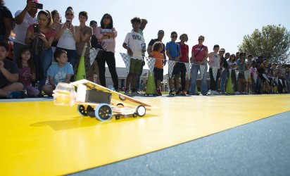 A line of onlookers watch and film as a small solar call shaped to a point races across a yellow track laid out over a blue turf, with triangular flags behind.
